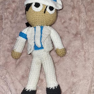 May include: Crocheted doll in a white suit, hat, and black shoes. The doll has a tan face, black hair, and large eyes. The hat has a blue band. The doll's jacket has a blue stripe on the arm and a blue stripe down the front.