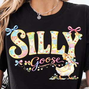 Puede incluir: Camiseta negra con las palabras "SILLY Goose" en un diseño de patchwork colorido. El diseño incluye un ganso de dibujos animados con sombrero y lazo, rodeado de flores y lazos.