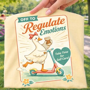 Pode incluir: Camiseta amarelo claro com um desenho de ganso de desenho animado em uma scooter, segurando uma xícara de café. O texto diz "Off to Regulate Emotions" e "Take Time for Self Care!" em estilo retrô.