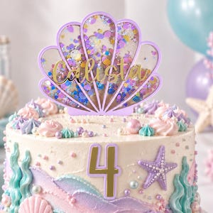 May include: A round cake decorated with pastel frosting, edible pearls, and sea-themed accents. A purple shell-shaped shaker topper filled with colorful sequins features the name Calista in gold script, and a gold number 4 is on the front.