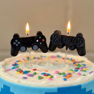 May include: A birthday cake with blue frosting and colorful sprinkles, topped with two black controller-shaped candles with lit flames. The cake has a white frosting layer and a blue pixelated design around the base.