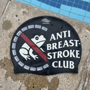 Può includere: Cuffia da nuoto nera con la scritta bianca "ANTI BREAST-STROKE CLUB". Una rana bianca è all'interno di un cerchio rosso con una linea che lo attraversa. Il marchio "cutwave" è stampato in alto.