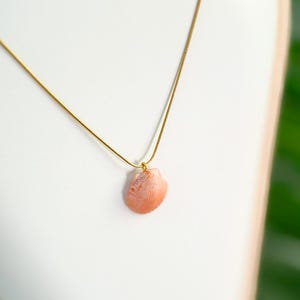 May include: A delicate gold-tone necklace featuring a small, peach-colored seashell pendant. The seashell has a textured surface and is suspended from a thin, gold chain. The necklace is displayed on a white background.