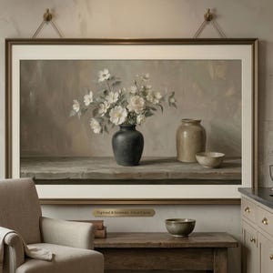 May include: A framed still-life painting depicts a dark vase filled with white flowers, a tan jar, and a small bowl on a wooden surface. The painting is in a room with a neutral color palette, a beige armchair, and a copper kettle.