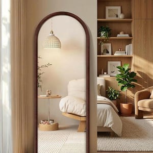 May include: A tall, arched-top mirror with a dark brown frame. The mirror reflects a bedroom scene with a bed, side table, and a woven pendant light. The room has a neutral color palette with wooden accents.