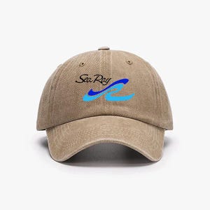Könnte beinhalten: Beige Baseball Cap mit dem Text "Sea Ray" in schwarzer Schrift und einem blau-hellblauen Wellenmuster. Die Kappe hat einen gebogenen Schirm und einen Knopf oben.