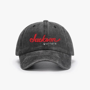 Puede incluir: Gorra de béisbol negra desgastada con el texto rojo "Jackson guitars" en cursiva. La gorra tiene una visera curva y un cierre de correa de tela. Accesorio musical.