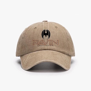 May include: A tan baseball cap with the text "RAVIN CROSSBOWS" in red and black. The cap features a black graphic of a stylized bat or bird. The hat has a curved brim and a fabric strap closure.
