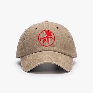 Puede incluir: Gorra de béisbol beige con visera curva. La gorra presenta un emblema circular rojo en la parte delantera con un diseño estilizado. La gorra está hecha de un material suave y duradero y tiene un estilo clásico e informal.