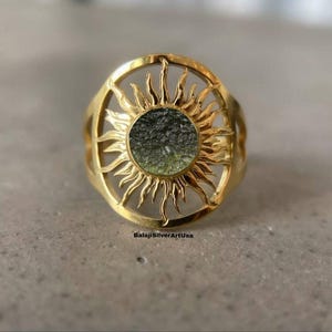 Könnte beinhalten: Ein goldfarbener Ring mit einem Sonnenmuster. Der Ring hat einen kreisförmigen, strukturierten, dunkelgrünen Stein in der Mitte. Das Sonnenmuster ist offen, mit nach außen gerichteten Strahlen. Der Text "BalajiSilverArtUsa" ist sichtbar.