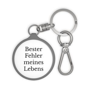 May include: A silver-coloured keychain with a round pendant. The pendant has a white background with the German text "Bester Fehler meines Lebens" in black. The keychain includes a split ring and a lobster clasp.
