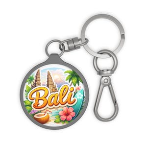 Puede incluir: Un llavero redondo de Bali con una escena de playa tropical. El diseño incluye una tabla de surf, palmeras, una bebida de coco y la palabra "Bali" en escritura naranja. El llavero tiene un anillo y un cierre plateados.