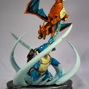 May include: A detailed figurine featuring two characters in a battle pose. The top figure is orange with blue wings, while the bottom figure is blue and yellow. The base is a dark gray color, and the figures are surrounded by light blue swirls.