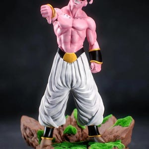 May include: A pink figurine of a muscular character with white trousers, a black belt with a gold buckle, and black wristbands. The character has a pointed head and is standing on a rocky base with green moss.