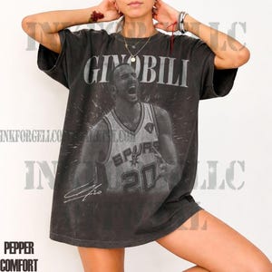 May include: Dark gray t-shirt featuring a black and white image of basketball player Ginobili. The image includes the name "GINOBILI" above the player and his signature below. The shirt has a vintage, distressed look.