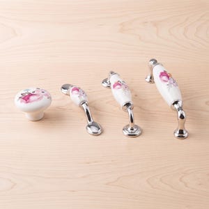 May include: A set of four cabinet hardware pieces including one round knob and three arched handles. Each piece is made of white ceramic with a floral pattern featuring pink and purple flowers, accented by polished silver-tone metal bases.