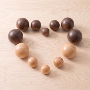 May include: A collection of eleven smooth, spherical wooden balls arranged in a heart shape. The set includes five dark brown walnut-toned spheres and six lighter tan-coloured spheres of varying sizes, all featuring a polished, natural wood grain finish.