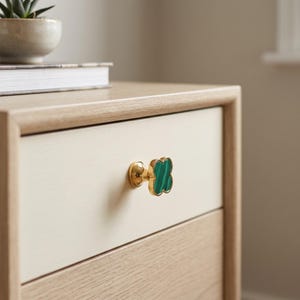 May include: A clover-shaped cabinet knob featuring a polished gold-tone metal base and a green malachite inlay with distinct dark green banding. The knob is attached to a light-colored wooden drawer front with a smooth, matte finish and clean edges.
