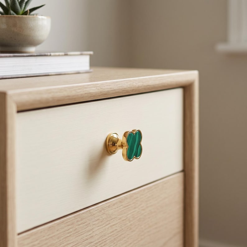 May include: A clover-shaped cabinet knob featuring a polished gold-tone metal base and a green malachite inlay with distinct dark green banding. The knob is attached to a light-colored wooden drawer front with a smooth, matte finish and clean edges.