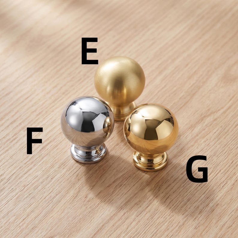 以下が含まれることがあります： E、F、Gとラベル付けされた3つの球状のキャビネットノブ。ノブEはマットゴールド仕上げ、ノブFはポリッシュクローム仕上げ、ノブGはポリッシュゴールド仕上げです。各ノブには円形のベースと滑らかで丸みを帯びた上面があります。