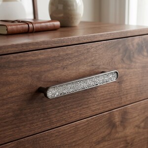 May include: A horizontal drawer pull handle featuring a dark metallic frame filled with a textured, sparkling clear crystal inlay, mounted onto a dark brown wood grain drawer front with a smooth, polished finish and visible wood texture.