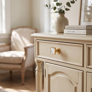 May include: A cream-colored wooden cabinet featuring a single drawer with a gold-rimmed, four-leaf clover-shaped knob with a white pearlescent inlay. The drawer front is framed by molding, and the cabinet has a smooth, light-toned finish.