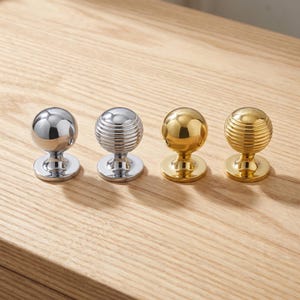 Peut inclure: Quatre boutons de placard alignés. Deux boutons sont argentés, l'un avec une finition lisse et l'autre avec une texture nervurée. Deux boutons sont dorés, l'un avec une finition lisse et l'autre avec une texture nervurée. Chaque bouton a une base circulaire.