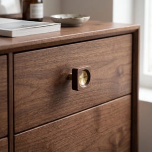 May include: A square-shaped cabinet knob featuring a dark brown wooden frame with a circular, polished brass centre inlay. The knob is mounted on a dark brown wooden drawer front with a visible wood grain pattern, showcasing a smooth, finished surface.