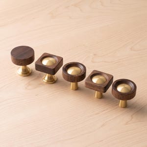 May include: A set of five cabinet knobs featuring dark brown wood and polished brass metal components. The collection includes circular and square wooden bases, each centred with a smooth, rounded brass inlay and supported by a brass pedestal.
