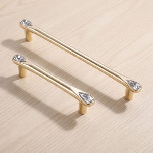 Peut inclure: Deux poignées de placard en métal doré de longueurs différentes, chacune présentant une barre cylindrique lisse. Les deux extrémités de chaque poignée sont ornées d'une incrustation de cristal transparent en forme de goutte.
