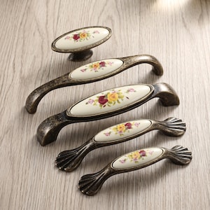May include: A collection of five cabinet hardware pieces featuring an oval-shaped white ceramic center with a floral pattern of red and yellow roses. The hardware has a dark, distressed bronze-colored metal finish with curved handles.