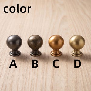 以下が含まれることがあります： A、B、C、Dとラベル付けされた4つの球状のキャビネットノブ。ノブAはダークチャコール、Bはアンティークブロンズ、Cは温かみのある銅色、Dはマットゴールドです。各ノブは滑らかで丸みを帯びたトップと円形のベースを備えており、一列に並んでいます。