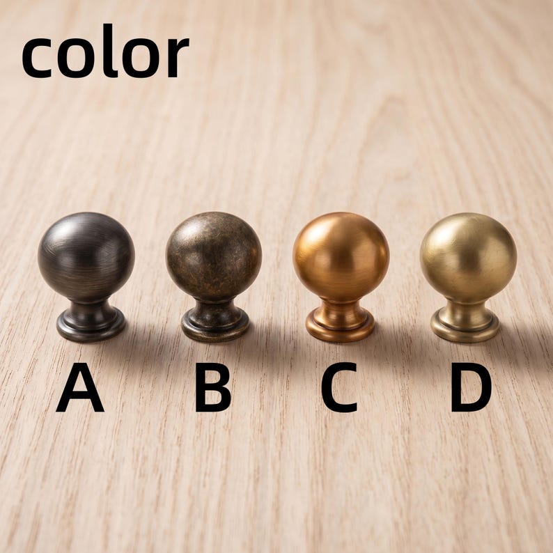 以下が含まれることがあります： A、B、C、Dとラベル付けされた4つの球状のキャビネットノブ。ノブAはダークチャコール、Bはアンティークブロンズ、Cは温かみのある銅色、Dはマットゴールドです。各ノブは滑らかで丸みを帯びたトップと円形のベースを備えており、一列に並んでいます。