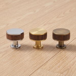 May include: Three cylindrical cabinet knobs featuring a dark brown wood center band. The left knob has a polished chrome finish, the center knob has a brushed gold finish, and the right knob has an antique brass finish with a circular top surface.
