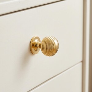 Pode incluir: Um puxador de m&oacute;vel em metal dourado polido com formato esf&eacute;rico e uma s&eacute;rie de ranhuras conc&ecirc;ntricas horizontais. O puxador est&aacute; montado numa placa de base circular e fixado numa frente de gaveta plana de cor branco-sujo.