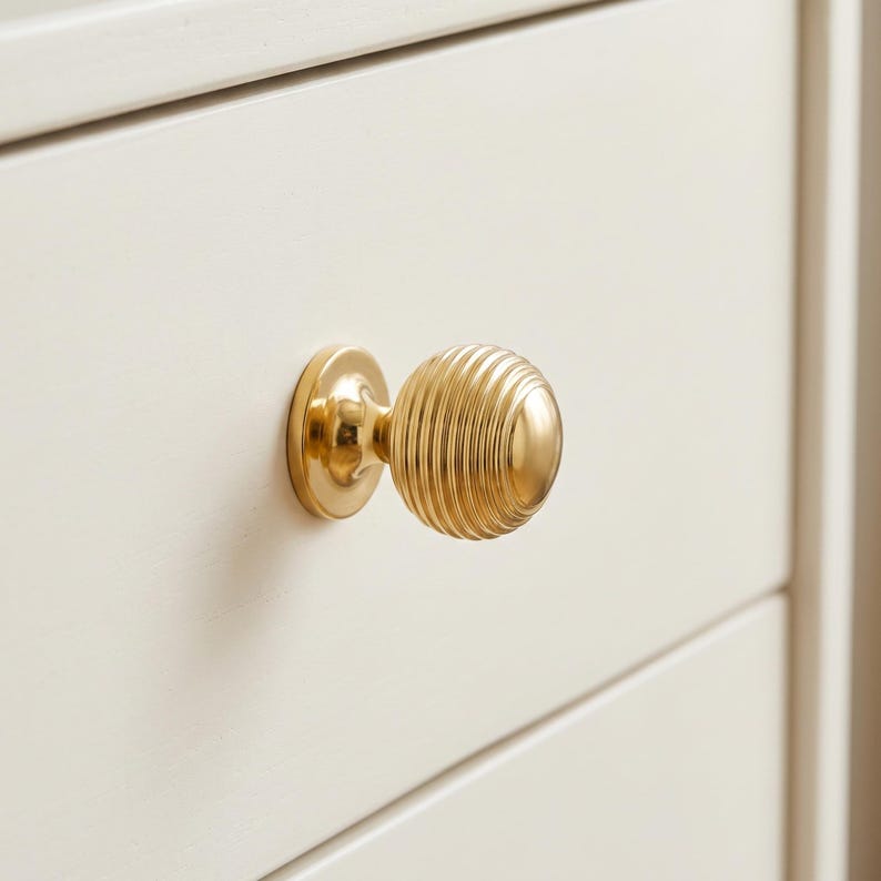 Pode incluir: Um puxador de m&oacute;vel em metal dourado polido com formato esf&eacute;rico e uma s&eacute;rie de ranhuras conc&ecirc;ntricas horizontais. O puxador est&aacute; montado numa placa de base circular e fixado numa frente de gaveta plana de cor branco-sujo.