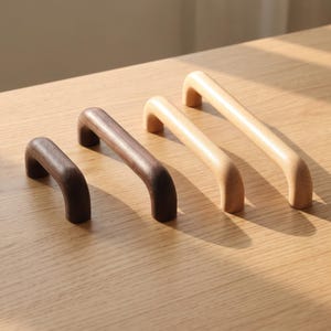 May include: Four wooden cabinet handles arranged in a row, featuring two dark brown walnut-toned handles and two light beige natural wood-toned handles. Each handle has a smooth, rounded arch shape with two mounting points for attachment to furniture.