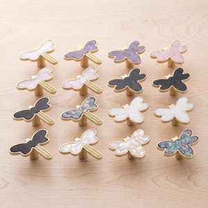 May include: A collection of sixteen dragonfly-shaped cabinet knobs arranged in a four-by-four grid. Each knob features a gold-toned metal base and a flat, decorative top surface in various colors and patterns, including white, black, purple, pink, and iridescent finishes.