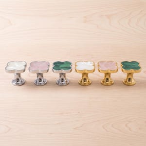 May include: A set of six clover-shaped cabinet knobs arranged in a row. Three knobs feature silver-toned bases with white, pink, and green marbled inlays. The other three knobs have gold-toned bases with white, pink, and green marbled inlays.