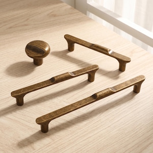 Op de afbeelding: Een set antiek messing deurbeslag bestaande uit één ronde knop en drie langwerpige ladehandgrepen. Elke handgreep heeft een glad, horizontaal ontwerp met een subtiele inkeping in het midden en twee cilindrische bevestigingspunten.