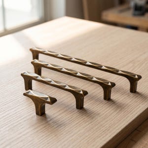 May include: Four brass-toned metal cabinet pulls of varying lengths arranged in a diagonal line. Each pull features a textured, faceted surface with a repeating geometric pattern along the top edge and two vertical mounting posts at the ends.