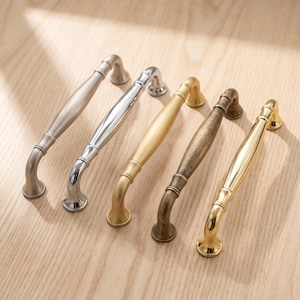 May include: Five metal drawer pulls arranged in a row, featuring a curved, elongated design with rounded ends. The finishes include brushed nickel, polished chrome, matte gold, antique bronze, and polished brass, each with a smooth, arched handle.