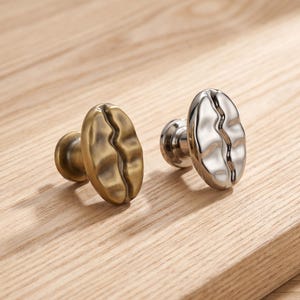 May include: Two cabinet knobs shaped like coffee beans. One knob has a matte antique brass finish, while the other features a polished chrome finish. Both knobs have a central vertical indentation and a circular base for mounting to furniture.