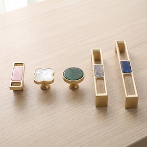 May include: A collection of five gold-toned metal cabinet hardware pieces. The set includes two rectangular pulls with pink and blue inserts, a clover-shaped knob with a white insert, a round knob with a green insert, and a rectangular pull with a gray insert.