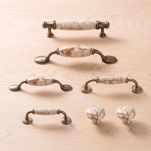 May include: A collection of cabinet hardware including four arched drawer pulls and two round knobs. Each piece features a white ceramic center with brown marble-like veining and antique brass metal ends or bases for mounting to furniture.