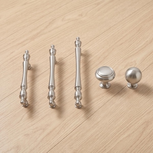 Può includere: Un set di cinque componenti di ferramenta per mobili con finitura in nichel spazzolato. Il set comprende tre maniglie per cassetti allungate e sottili con dettagli arrotondati decorativi e due pomelli circolari, uno piatto e uno sferico.