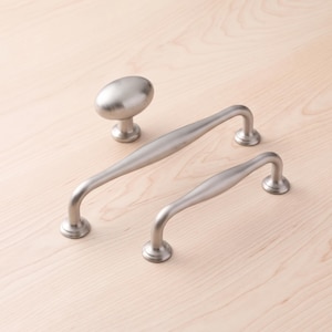 Può includere: Un set di tre componenti di ferramenta composto da un pomello per mobile di forma ovale e due maniglie ad arco. Ogni elemento presenta una finitura in nichel satinato spazzolato color argento metallizzato con basi di montaggio circolari.