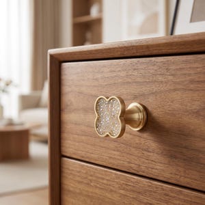May include: A clover-shaped drawer knob featuring a brushed gold metal frame and a textured, multifaceted clear crystal center. The knob is mounted on a wooden drawer front with a visible grain pattern, showcasing a metallic base and stem.