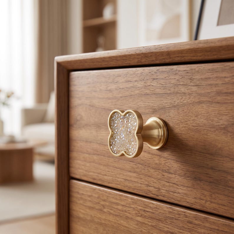 May include: A clover-shaped drawer knob featuring a brushed gold metal frame and a textured, multifaceted clear crystal center. The knob is mounted on a wooden drawer front with a visible grain pattern, showcasing a metallic base and stem.