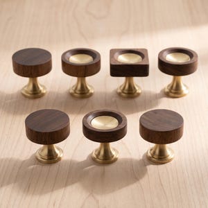Puede incluir: Una colección de siete pomos para armarios con tapas de madera marrón oscuro y bases de metal dorado. Los pomos incluyen formas circulares y cuadradas sólidas, así como diseños circulares con una incrustación central cóncava dorada.